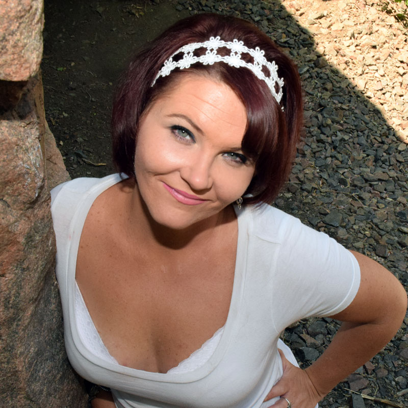 A smiling woman with short, dark red hair, wearing a white top with a delicate lace headband. She stands confidently outdoors, leaning against a stone wall with a relaxed and friendly expression, embodying both warmth and approachability.