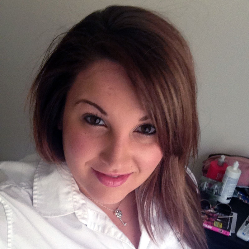 A young woman with long, brown hair, smiling warmly at the camera. She is wearing a crisp white blouse and a delicate necklace, exuding confidence and approachability. The setting appears to be indoors, with personal items visible in the background, adding a casual and relaxed atmosphere to the image.